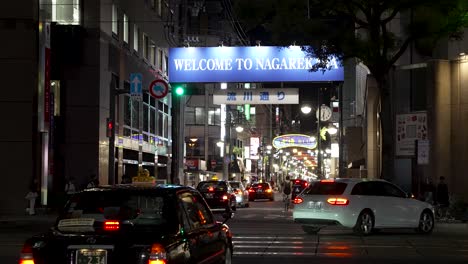 Willkommen-Beim-Nagarekawa-Plakatschild-Bei-Nacht-Mit-Darunter-Vorbeifahrendem-Verkehr-In-Hiroshima