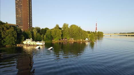 Wakeboard-Park-Mit-Einem-Alten-Schiff-Vor-Einem-Sowjetisch-Aussehenden-Fernsehgebäude-Mit-Einem-Fernsehturm-Im-Hintergrund-Am-Fluss-Daugava