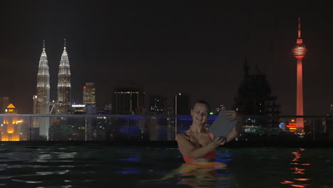 View-of-woman-in-swimming-pool-on-the-skyscraper-roof-making-selfie-by-tablet-against-night-city-landscape-Kuala-Lumpur-Malaysia