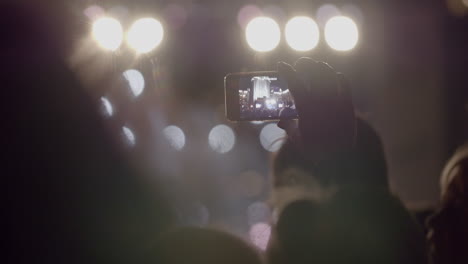 Spectator-man-recording-video-of-stage-in-front-of-bright-spotlights-via-smartphone-at-outdoor-music-concert