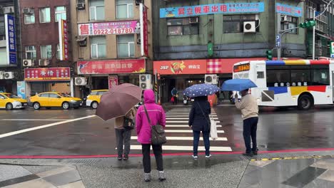 Pedestrians-carrying-umbrellas-waiting-at-the-traffic-light-to-turn-green-on-a-rainy-day-in-Keelung-City,-Ren’ai-District,-Taiwan