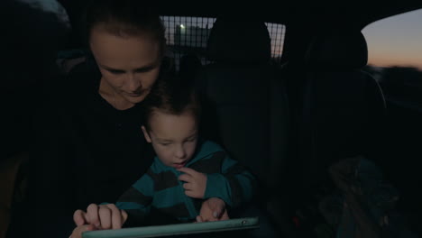 View-of-woman-with-son-in-the-dark-riding-in-car-on-back-seat-watching-computer-tablet