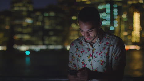 Man-Using-Mobile-Phone-At-Night-With-City-Skyline-In-Background