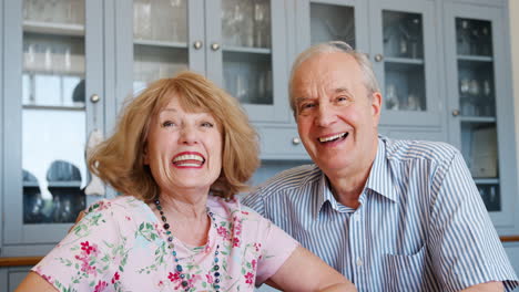 Portrait-Of-Smiling-Senior-Couple-Sitting-At-Kitchen-Table