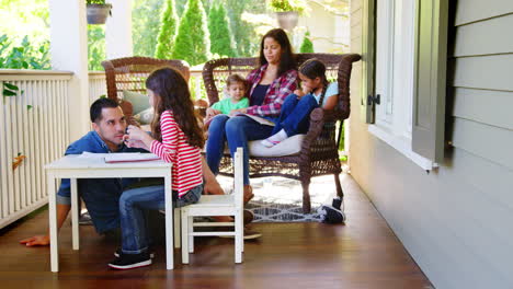 Family-Sit-On-Porch-Of-House-Reading-Books-And-Playing-Games