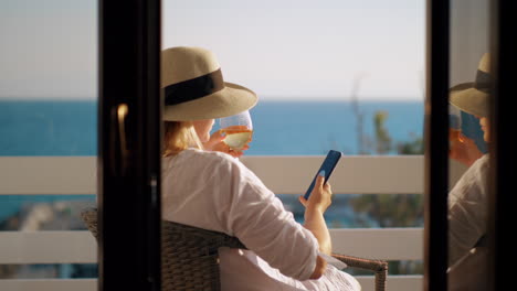 Frau-Mit-Weinglas-Nutzt-Handy-Beim-Entspannen-Auf-Dem-Balkon-Mit-Blick-Auf-Das-Meer