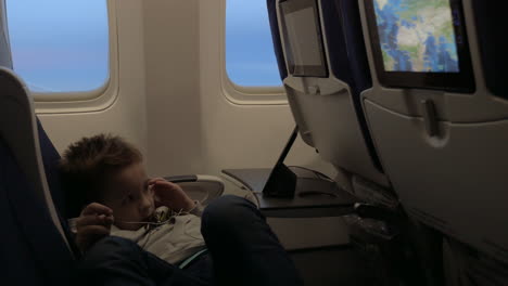 Blick-Auf-Einen-Kleinen-Jungen,-Der-Sich-Im-Flugzeug-Filme-Im-Headset-Ansieht-Und-Auf-Dem-Sitz-Vor-Dem-Flugzeugfenster-Liegt