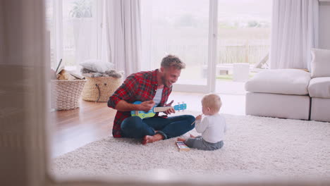 Dad-sits-playing-ukulele-with-toddler,-seen-through-window