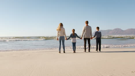 Rear-View-Of-Family-On-Winter-Beach-Holding-Hands-Looking-At-Sea