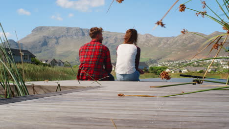 Couple-sitting-on-a-jetty-by-a-lake-and-mountains,-back-view