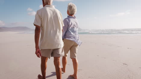 Rear-View-Of-Romantic-Senior-Couple-On-Summer-Vacation-Walking-Along-Beach