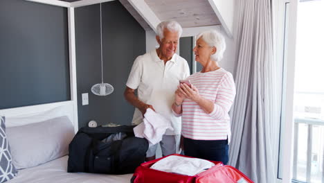 Älteres-Paar-Im-Schlafzimmer-Schaut-Aufs-Handy,-Während-Es-Koffer-Für-Den-Urlaub-Packt