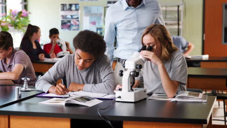 Estudiantes-De-Secundaria-Con-Tutor-Usando-Microscopio-En-Clase-De-Biología