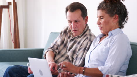 Senior-Couple-Sitting-On-Sofa-At-Home-Using-Laptop-Computer-To-Shop-Online