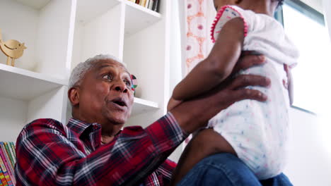 Grandfather-Lifting-Baby-Granddaughter-Into-The-Air-And-Smiling-At-Home