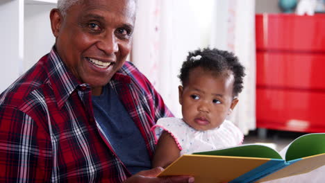 Grandfather-Sitting-And-Reading-Book-With-Baby-Granddaughter-At-Home