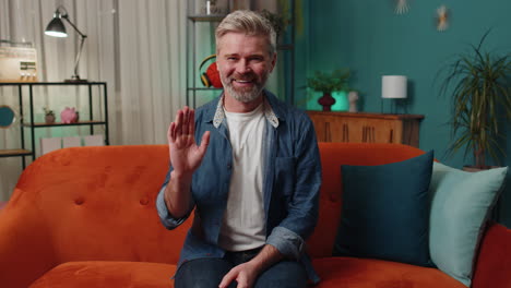 Mature-man-smiling-friendly-at-camera,-waving-hands-gesturing-hello,-hi,-greeting-at-home-on-couch