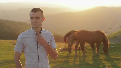 Young-man-is-standing-in-the-field-near-mountains.-Behind-him-grazing-horses