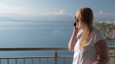 A-fair-haired-woman-talking-to-a-phone-on-a-hotel-balcony-near-the-sea-landscape