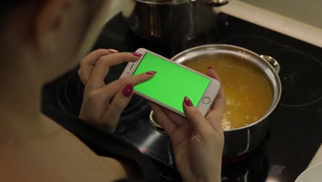 Woman-is-holding-a-smartphone-with-a-green-screen,-and-cooking-soup
