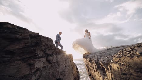 Groom-goes-to-bride-and-gives-her-a-hand.-Newlyweds-on-mountainside-by-the-sea