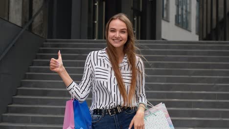Trendy-girl-holding-shopping-bags-and-showing-thumbs-up.-Rejoicing-with-good-holiday-sale-discounts
