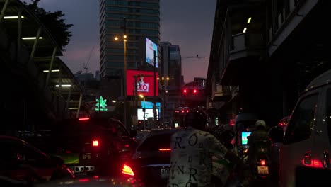 Heavy-traffic-stops-at-a-crossroad-in-Bangkok-revealing-all-kinds-of-motorists-while-billboards-playing-and-a-Marijuana-neon-sign,-Thailand