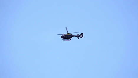 En-Cámara-Lenta,-Un-Helicóptero-De-Policía-G-polf-Airbus-Ec135t2-De-Doble-Rotor-Negro-Y-Amarillo-Se-Cierne-Sobre-Un-Cielo-Azul-Claro