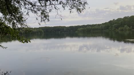 Kleiner-See-In-Der-Nähe-Von-Köln-Mit-Ruhigem-Wasser-Mit-Bäumen-Rundherum-Und-Am-Ufer-Unter-Einem-Bewölkten-Himmel-Mit-Wolken