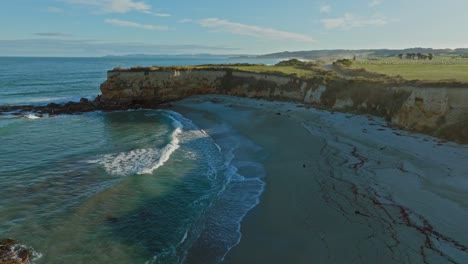 Beautiful-aerial-view-of-Mitchells-Rocks-with-secluded,-sandy-beach,-crumbling-coastal-cliffs-and-ocean-spray-over-coastal-landscape-in-Otago,-South-Island-of-New-Zealand-Aotearoa