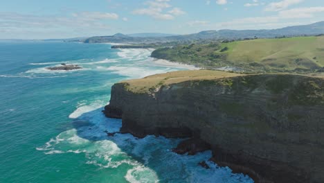 Aerial-view-of-steep,-rugged-and-dramatic-cliffs-and-wild,-rough-ocean-with-white-wash-waves-at-Smails-Beach-on-the-Otago-Peninsula,-Dunedin,-New-Zealand-Aotearoa