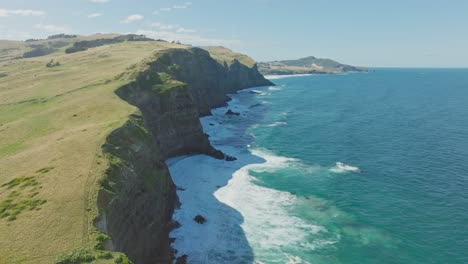 Dramatic-aerial-view-over-steep-rocky-cliffs-and-white-wash-ocean-waves-at-Smails-Beach-on-the-Otago-Peninsula,-Dunedin,-New-Zealand-Aotearoa