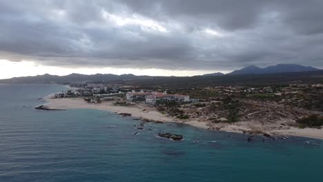Aerial-view-over-the-beautiful-coast-of-El-Chileno-Beach-baja-california-sur,-Mexico-with-a-view-of-the-blue-sea,-hotel-buildings-and-landscape-with-lush-vegetation-with-magnificent-mountain-ranges