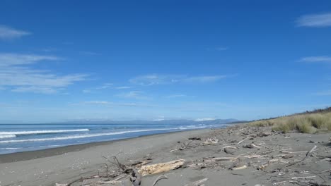 Small-waves-roll-into-driftwood-strewn-sandy-beach-on-a-sunny-winter's-day-with-snow-visible-on-distant-hills---Pegasus-Bay,-New-Zealand