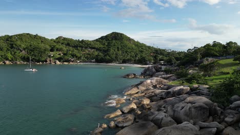 Static-shot-of-a-rocky-shore-near-the-mountains-with-still-waters-and-a-boat-on-the-sea-Koh-Samui-islands