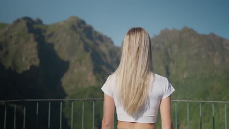 Blond-fit-woman-walking-towards-steel-railing-at-mountain-viewpoint