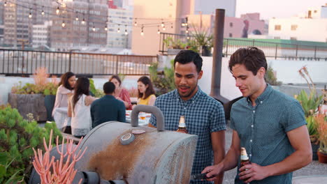 Two-Men-Cooking-Barbecue-For-Friends-Gathered-On-Rooftop-Terrace-With-City-Skyline-In-Background
