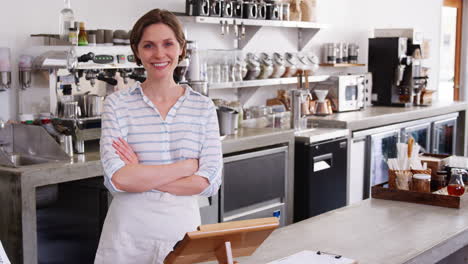 Young-woman-behind-counter-at-a-coffee-shop-smiles-to-camera