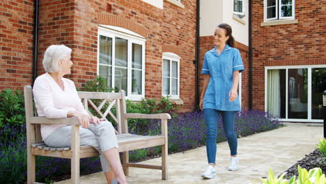 Senior-Woman-Sitting-On-Bench-And-Talking-With-Nurse-In-Retirement-Home