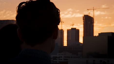 Rear-View-Of-Couple-On-Rooftop-Terrace-Looking-Out-Over-City-Skyline-At-Sunset
