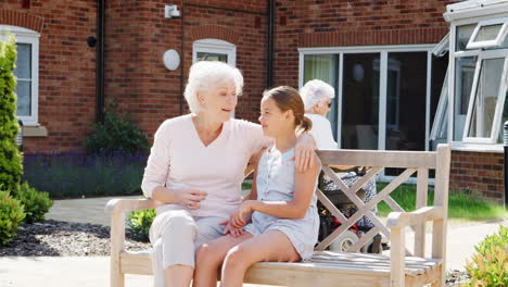 Granddaughter-Sitting-On-Bench-With-Grandmother-During-Visit-To-Retirement-Home