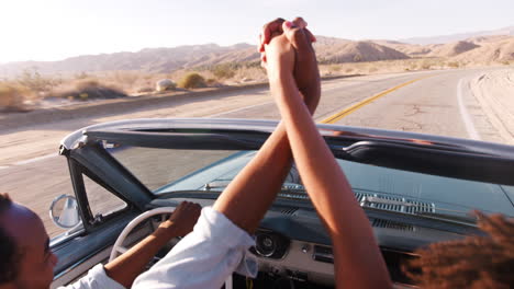 Couple-in-open-top-car-hold-hands-in-the-air,-elevated-view