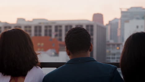 Rear-View-Of-Friends-Gathered-On-Rooftop-Terrace-Looking-Out-Over-City-Skyline