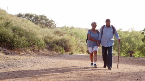 Senior-Couple-Wearing-Backpacks-Hiking-In-Countryside-Together