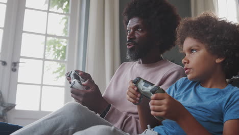 Happy-middle-aged-black-father-and-his-pre-teen-son-sitting-on-the-floor-in-their-living-room-talking-and-playing-video-games-together,-close-up
