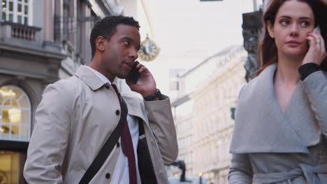 Young-black-businessman-standing-on-street-talking-on-smartphone,-white-millennial-woman-walking-past-also-using-phone,-close-up,-low-angle