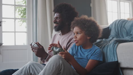 Black-middle-aged-father-and-his-pre-teen-son-sitting-on-the-floor-in-their-living-room-playing-video-games-together,-close-up