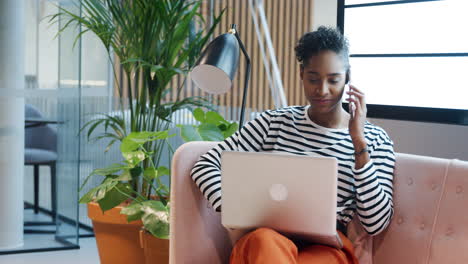 Young-adult-mixed-race-businesswoman-sitting-on-a-sofa-in-a-casual-office-using-phone-while-working-on-a-laptop-computer-balanced-on-her-knees
