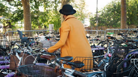 Young-woman-wearing-unbuttoned-yellow-pea-coat,-black-hat-and-sunglasses-parking-her-bicycle,-putting-on-crossbody-bag-and-walking-away-smiling,-side-view,-tracking-shot