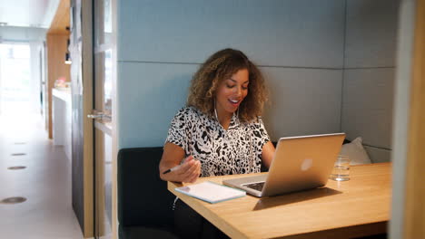 Close-up-of-young-mixed-race-woman-working-alone-on-a-laptop-at-a-table-in-a-booth-at-an-office-canteen-talking-and-smiling,-zoom-in-shot